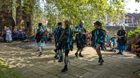 Bradford-on-Avon Town Council Several people wearing black and blue matching outfits, eye masks, and some with black hats, dancing on a brick path with a large tree in the background. There are some people watching , including a group of women in blue dresses.