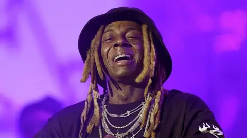 Getty Images Um homem, sorrindo no palco, usando um chapéu, vários colares de prata e uma blusa preta. O fundo é uma névoa roxa.