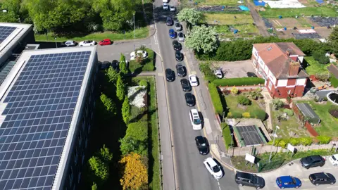 PA Media Aerial view of about 15 cars queuing to get into a household recycling centre in Birmingham. Other cars stretch into the distance. On the right is what seems a house and garden, with an industrial building opposite it on the left, across a road.