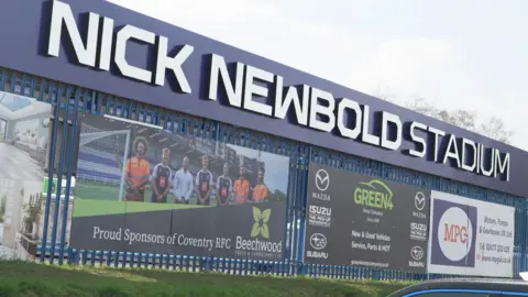 The Nick Newbold Stadium sign is in silver letters on a blue board with advertising banners on display underneath it.