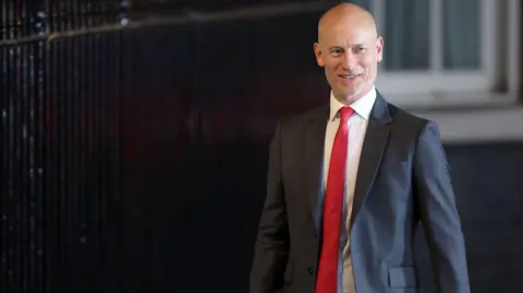 Getty Images A bald man in a dark suit, white shirt and red tie. 