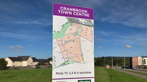 A sign with writing at the top stating "Cranbrook Town Centre" in white writing on a purple background. Below is a map of Cranbrook, with writing saying 'Plots TC 2,3 & 4 available". In the background are new white houses and a large green space. There is a road to the right, with another brown and white building in the distance.