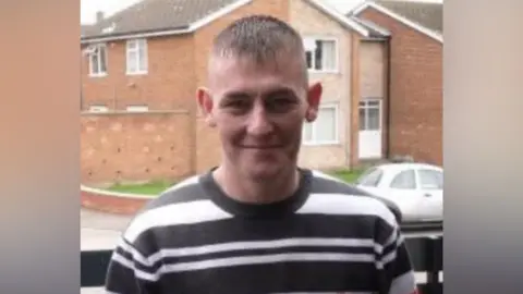 David Cuthbert. He is smiling at the camera and has short hair which has been shaved at the sides. He is wearing a black and white striped top.
