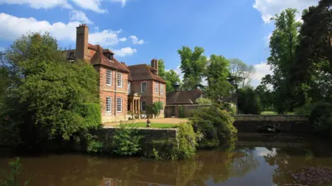 An image of Groombridge Place, near Tunbridge Wells. The sun in shining and there is water in front of a large manor house. 