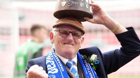 PA Media Oldham Athletic owner Frank Rothwell celebrates promotion to the EFL following the Vanarama National League play-off final at Wembley Stadium. He is wearing a blue and white Oldham Athletic scarf over his navy blue suit and has a trophy resting on his head. He has a fist up celebrating the club's promotion.