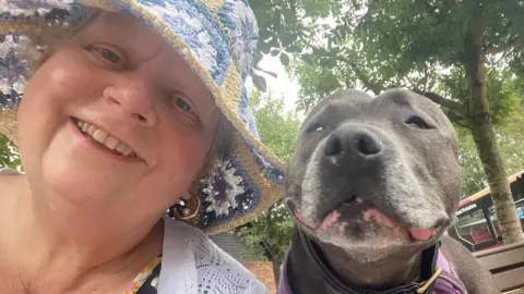 A selfie-style photograph of a woman smiling while wearing a hat. A dog is next to her face and looks towards the camera. There are trees in the background.