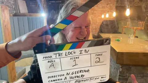 Simon Thake A clapperboard is held up infront of the face of a man with long white hair and gritted teeth