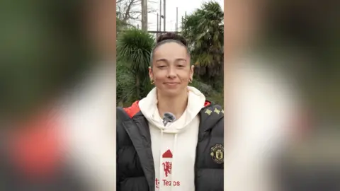 Phallon Tullis-Joyce stands in front of trees and smiles at the camera. She wears a white hoodie under a black puffer jacket.