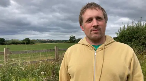 BBC Councillor Andrew Matthews wearing a yellow hoodie and looking into the camera. He is standing in front of a field and fence under a grey sky