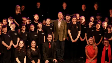 Viva Arts and Community Group Prince Edward is wearing a suit and is surrounded by young people and adults, all wearing black except for one person in red, inside a dark theatre room.