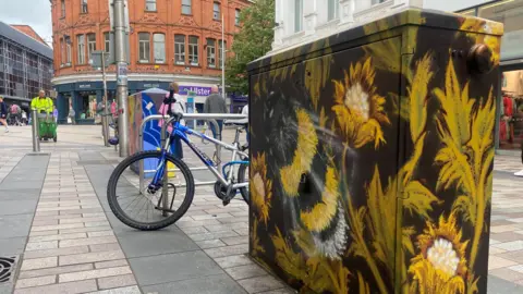 A utility box in Belfast city centre which has sunflowers painted on it. There is a street and shops in the background.