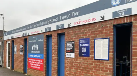 Emma Baugh/BBC Outdoor view of the turnstile building. It is red bricked with a sign over the door directing ticket-holders though the turnstiles. There is a sign on the wall reading 'Home fans only'. There are five thin blue doors for fans to push through to enter the stadium. 