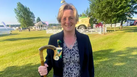 BBC/ Cara Thorpe A woman stands in front of a tractor and holds a staff which reads "show director".