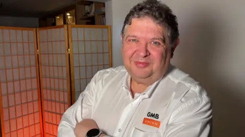 Ben Schofield/BBC Steve Garelick standing inside, with his arms folded, and looking directly down the camera. He is wearing a white, open necked shirt, with GMB Union embroidered on the chest. He has curly dark hair, flecked with grey, and worn relatively short. Behind him on the right of the image is a blank white wall, while on the left is a decorative screen, lit orange from below.