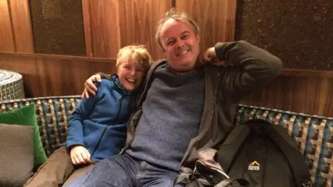 A man and a boy are seated closely together on a patterned bench in an indoor setting with wood-panelled walls. The boy on the left wears a blue jacket, while the man on the right wears a dark sweater and jacket. A backpack rests beside them.