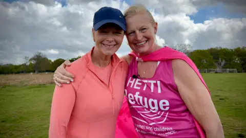 Hetti Barkworth-Nanton is pictured with her friend in a field. They are both smiling and looking directly at the camera. Hetti is on the right and is wearing a pink bib and cape. She has her arm around her friend who is to the left of her. 