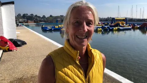 Research patient Lynne MacFarlane smiling for the camera by a pool. She is wearing a yellow vest. There are some swimming aids in the water.