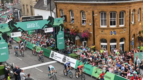 SWpix.com Cyclists riding over the finish line in a town with crowds cheering behind the hoardings. The finish line has a green board with "West Northamptonshire" written on the top.