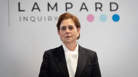 Baroness Kate Lampard wears a black jacket over a white shirt. She stands in front of a white background with the wording "Lampard Inquiry" in black letters, followed by three coloured dots.