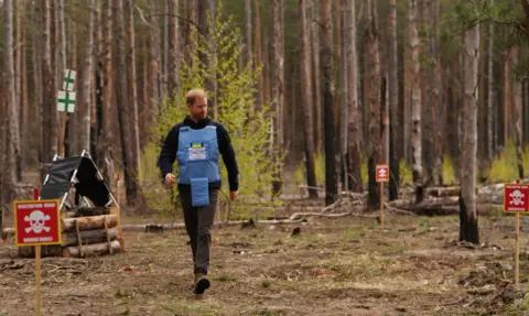 Prince Harry, a white man with short red hair, walking through a woodland in a blue vest and grey trousers. He is walking past signs that have a skull and bones image on them and writing. 