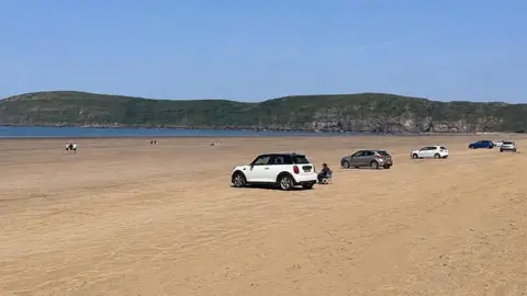 A row of five cars parked on the sand at Brean beach, with a long green headland in the distance.