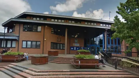 Google The St Albans City Council building. It is at least three storeys high and in front of it are some steps. A woman is sitting on a bench on the left in a flat area between to levels of steps, and a man is sitting on a bench on the right hand side. The building is red brick with windows and there is a tree on the right hand side.
