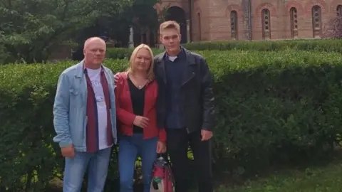 Illia Mitiushnikov Illia with his mum and dad. He is wearing a black leather jacket, his mum has shoulder-length blonde hair and is wearing blue jeans and a red jacket and his dad has short fair hair and is wearing a denim short and jeans. They are standing in front of a green bush with an old building behind them. 