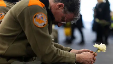 A bespectacled member of the civil protection unit wearing a khaki uniform, squats as he holds a white rose with both hands, with his head lowered 