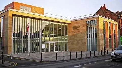View of the exterior of Cumbria House, the headquarters of Cumberland Council. The two-storey building has floor to ceiling windows and is made from sand-coloured brick. There is a precinct in front of it with bollards around the kerb.