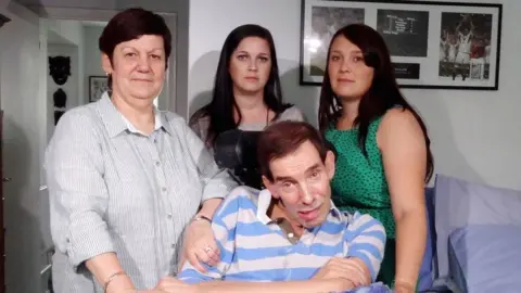 PA Media Tony Nicklinson sitting in his living room, wearing a white and blue striped shirt with his arms crossed. He is surrounded by three of his family members, who are looking at the camera with a solemn expression. 