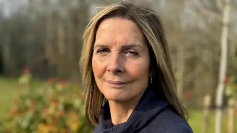 Jane Roche pictured with brown hair with blonde highlights in it. She is smiling slightly and wearing a blue roll-neck jumper.