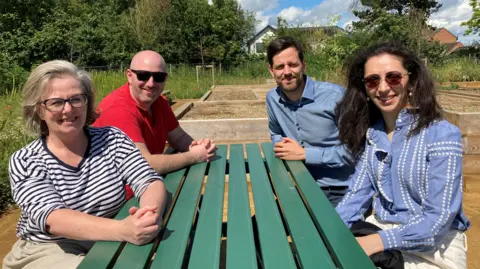 Two men and two women sit at a green picnic bench. In the background are raised beds, wild flowers and trees.