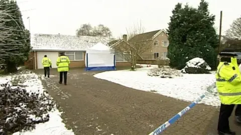 A snow-covered bungalow in Wisbech, where Una Crown lived. There is police tape across the drive and police officers on guard outside the tape and walking up to the house.