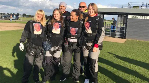 Family Breon's mother Kelly Simms with family and friends after a charity skydive in memory of her son. She is in the middle. Everyone has sky-dive gear on and t-shirts baring Breon's face. It is a bright and sunny day. They are standing on a patch of grass.