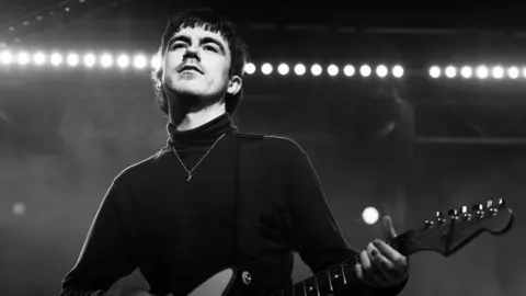 Lauren Skinner Declan McKenna wears a black polo neck and plays guitar on stage.