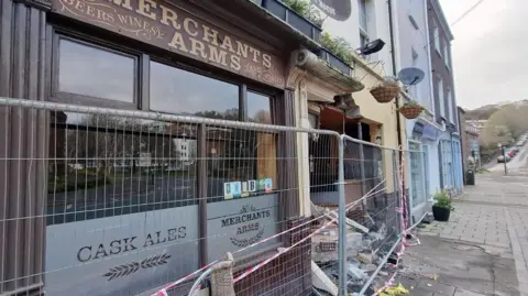 The outside of a pub after a car collided with it. There is a metal fence blocking the entrance, and there is debris in front of the building.