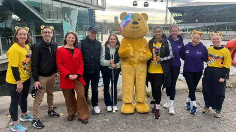 A big yellow bear puts his thumbs up while standing in the middle of a group of people by London's cable car, with the pods in the background. Eleanor is with her dad, Tim and is holding her white cane.