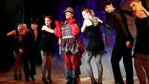 Barry Rivett Hotshots Photography Nigel Harman shows him in a black and red Fleshcreep costume. His shiny black waistcoat has red braiding and he wears a red shirt underneath with wide sleeves. His top hat style hat is black with red braiding. His hands are on his shoulders in a dance move and he is smiling. He has three dancers on each side of him wearing black costumes with red devil horns.