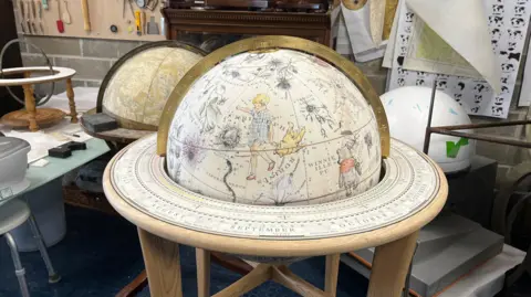 Tony Fisher/BBC A decorative globe sits on a wooden stand in a workshop, surrounded by tools, materials, and other partially assembled globes. The globe’s surface features intricate illustrations of plants, animals, and constellation-like figures, along with detailed labels. A brass meridian ring arcs over the top, while a circular calendar base beneath shows months such as “August,” “September,” and “October.” In the background, a wall displays neatly arranged tools, books, and reference materials used in crafting and designing globes.