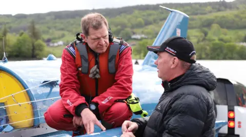 Matthew Willis DJ Gibbs crouches on Bluebird's right sponson as he chats to Dave Warby.
Gibbs has brown hair and is wearing a red pilot's outfit. Warby is wearing a black baseball cap and black jacket.
