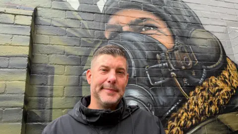 Steve Crowe is wearing a black jacket and is smiling. He is standing in front of a wall painting showing a World War Two airman wearing flying jacket, helment and oxygen mask.