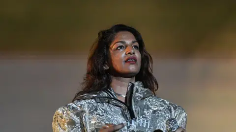 The artist is wearing a shiny silver jacket. She has long wavy dark brown hair and brown eyes. She is wearing red lipstick and looking off into the top right corner of the camera. 