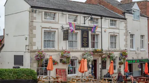 Graham Mitchell A three-storey white-coloured traditional pub with The Bailey Head on a sign above the entrance. Tables and benches are placed outside with people sitting on some of them.