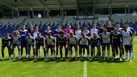 Glenavon FC Glenavon FC and Erzgebirge Aue line out before kick off. They are standing in the middle of the football pitch in front of a large stand inside the stadium. There are empty white and purple seats behind them. The Erzgebirge Aue players are wearing purple and white tops and Glenavon are wearing white and blue tops. Half of the players are standing upright and the others and leaning forward.