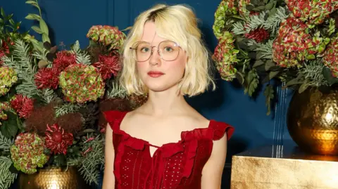 Maisie Peters wears a red dress with frills on the front and tie-front. She has short curly blonde hair and glasses and stands against a floral background