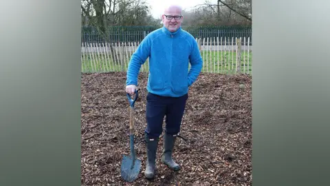 Lancashire County Council Councillor Shaun Turner wearing a blue fleece and navy trousers and wellies holding a spade resting on the ground in a park where new trees are being planted