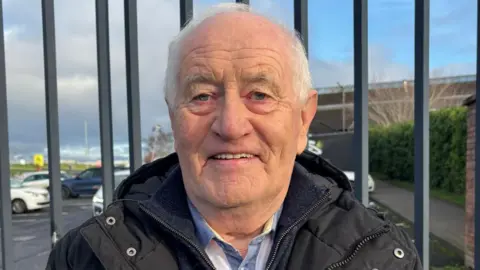 An older man wearing a black jacket stands smiling in front of a metal fence, with a car park and cloudy blue sky visible behind him.