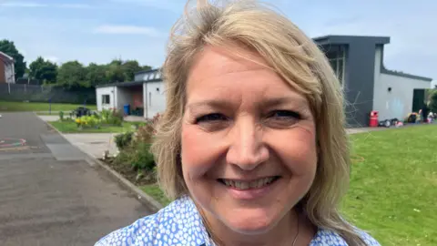 Rebecca is smiling. She has shoulder length blond hair and is wearing a blue blouse with a white pattern. She's standing outside.