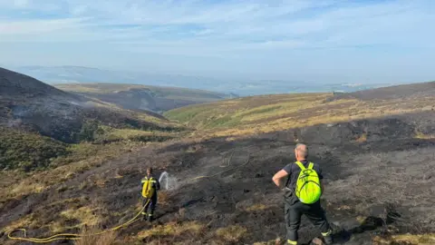 Two firefighters using a hose on a blackened area of moorland