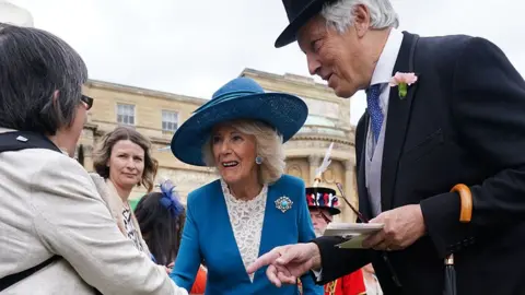 PA Media Queen Camilla wearing a blue dress and hat with lace inset, shakes hands with Jeanette Binns at the garden party as a man in dark top hat and tails introduces them